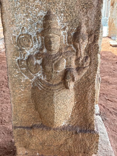 A close-up shot depicts an aged stone carving, likely a lintel or structural element, found in Kamalapura, India. The carving presents a stylized, reclining figure with multiple limbs, possibly a deity or mythical creature. The figure is situated amidst decorative carvings and appears weathered. The stone exhibits a rough texture with visible erosion and lichen growth. The background includes a blurred wall of similarly textured stone, emphasizing the age and historical context of the carving.