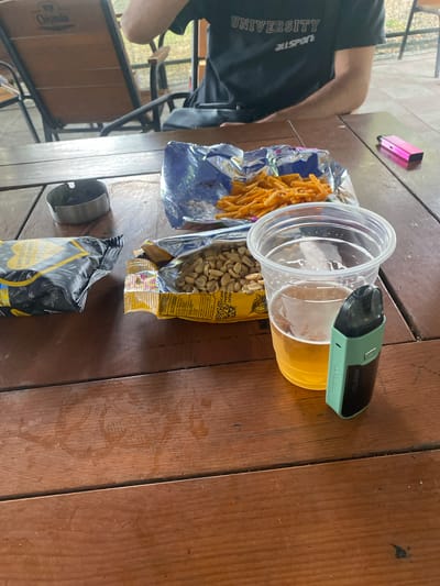 A partially visible person wearing a black t-shirt with "UNIVERSITY" and "ALLSPORT" printed on it sits at a wooden table outdoors. On the table are a disposable plastic cup filled with a light-colored beverage, a light green vape device, an opened bag of peanuts, an opened bag of orange puffed snacks, and a silver ashtray with ash. A pink electronic cigarette is visible on the table to the right. The scene is set at an outdoor dining area, possibly a restaurant or cafe. The overall impression is one of casual relaxation. The location is identified as Corjova, Moldova.