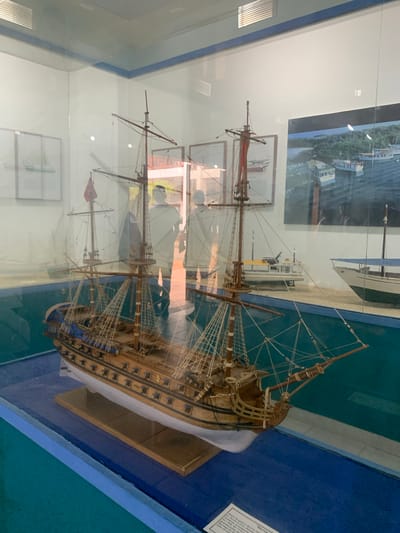The image shows a museum exhibit featuring a detailed wooden model of a historic sailing ship. The model is displayed in a glass case with a blue base that simulates water. The ship has multiple masts, sails, and rigging, with a red flag flying from the main mast. In the background, there are framed pictures of other vessels and a diorama of a harbor scene. Two faint silhouettes of people are visible through the glass, suggesting they are museum visitors. The environment appears to be indoors, likely in a museum located in Boca del Río, Venezuela, as indicated by the prompt. The lighting suggests it is daytime, and the reflections on the glass indicate the presence of overhead lights and the surrounding museum environment.