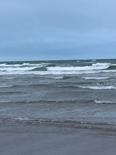 The image depicts a wide view of a choppy sea with several rows of waves breaking. The sky above is overcast and a pale blue-gray, suggesting a cloudy or misty day. The water is a dark, grayish-blue, reflecting the dullness of the sky. In the foreground, a dark, wet sand or shingle beach is visible, with the water lapping at its edge. The scene is devoid of any human presence or man-made objects, emphasizing the natural and somewhat desolate environment. The location is identified as Morfa Bychan, United Kingdom, a coastal area known for its beaches and natural landscapes.