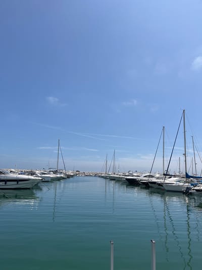 The image shows a marina in Nueva Andalucía, Spain. Several sailboats with masts are docked along a row. The water is calm and reflects the boats and sky. The sky is blue with few clouds, and contrails. Two grey steel poles are to the right of the frame.