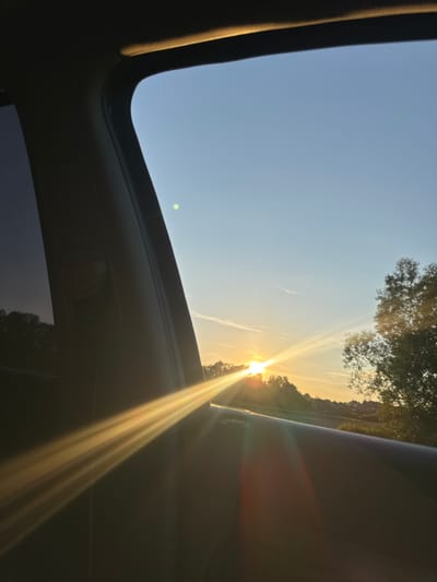 The image depicts a view through a car window at sunset. The sun is low on the horizon, casting golden light that streams across the scene. The sky is a clear blue with a few wispy clouds. Silhouetted trees line the horizon, with a hint of a treeline further in the distance. The foreground is dominated by the interior of the car, with the dark A-pillar and door panel visible. There is a distinct lens flare from the setting sun. The image does not provide any specific clues to suggest it was taken in Tovstoluh, Ukraine.