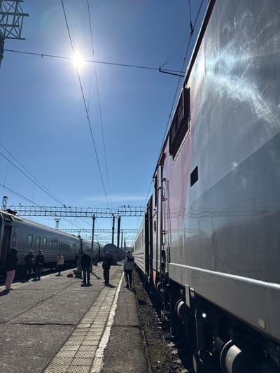 The image displays a train station platform in Kropachyovo, Russia, illuminated by direct sunlight under a clear, bright blue sky. Overhead, a complex network of electrical catenary lines and supporting gantries spans across multiple railway tracks.
On the immediate right, a large train car, presenting a two-tone metallic silver-grey and maroon-red exterior, is stationary. Its reflective metallic surface exhibits significant sun glare and visible circular polishing marks. Multiple vents and access hatches are discernable along its side.
To the left, on an adjacent track, another passenger train, characterized by its silver-grey color and numerous windows, is also stationary.
Between these two trains, on the concrete platform, approximately 10-15 individuals are observed. They are dressed in casual attire and are largely standing in small groups or individually. One person on the far left wears light pink pants. In the central foreground, near a cluster of people, items resembling luggage or personal belongings are situated on the platform. A solitary individual stands near the right-side train, facing away from the camera, possibly near an open doorway or car coupling. The platform surface is concrete, featuring tactile paving strips.