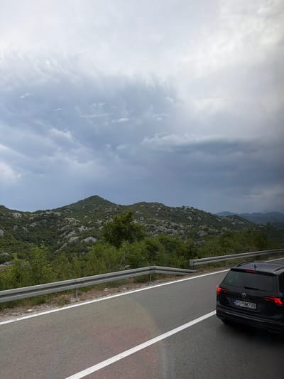 A black Volkswagen Tiguan is parked on the side of a road, its rear end facing the camera. The car has a license plate that reads "PG NM 799". The road is paved and has white lane markings. A metal guardrail runs along the edge of the road.
To the left of the road, a steep, rocky hillside is covered in dense green vegetation. The hillside stretches up towards a cloudy sky. The clouds are a mix of white and dark gray, suggesting overcast or stormy weather. The image appears to be taken during the day, with diffused light indicating it might be an overcast day or close to dusk/dawn.
The scene is set in a mountainous, possibly rural, location, with the vegetation and rocky terrain characteristic of such environments. There are no people visible in the image. The overall mood is calm and perhaps a bit dramatic due to the cloudy sky and rugged landscape. The text "TIGUAN" is visible on the rear of the car.