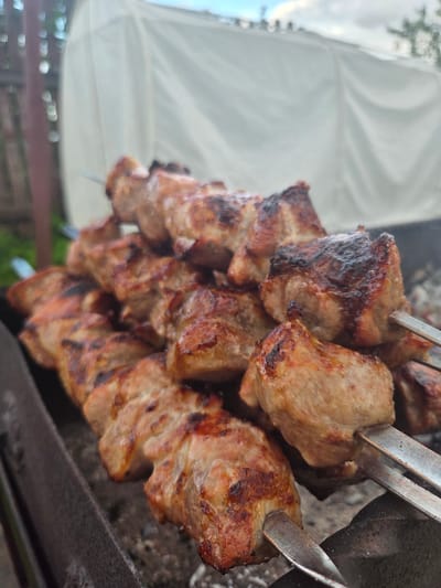 The image displays skewers of cooked meat, likely shashlik, arranged on metal skewers. The meat is a golden brown with some charring, indicating it has been grilled or roasted. The skewers are densely packed with chunks of meat, suggesting a generous portion. The background is blurred, but hints of an outdoor setting are visible. On the left, there is a white, tent-like structure, possibly an awning or canopy, which might suggest an outdoor event or gathering. Below this, there are hints of greenery and possibly a wooden fence or railing, further reinforcing the outdoor location. The lighting suggests it is daytime, possibly late afternoon or early evening, given the warm tones of the meat and the way the light falls. The overall impression is one of a casual, outdoor meal, perhaps a barbecue or picnic. There is no visible text in the image. The setting appears to be in Frolovo, Russia, as indicated by the provided context.