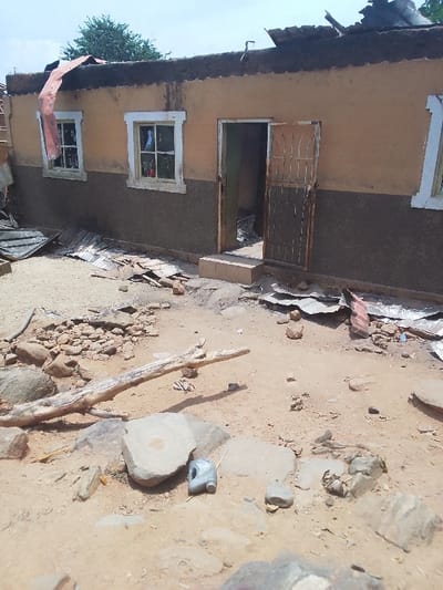 The image shows the exterior of a building in Kwal, Nigeria. The building is damaged, with its roof partially collapsed and debris scattered on the ground. The building is made of concrete blocks and has a brown facade. The roof is made of corrugated metal sheets. There are two windows and one doorway, with a metal gate. The windows have glass panes, and the doorway is open. The area surrounding the building is dusty and littered with debris. There is a large, broken tree branch lying on the ground. A blue plastic container is lying on the ground near the broken branch. The scene suggests that the building has been damaged by a storm or other natural disaster. There are no people present. The coordinates 9.804, 8.646 place this image in the city of Kwal, Nigeria.