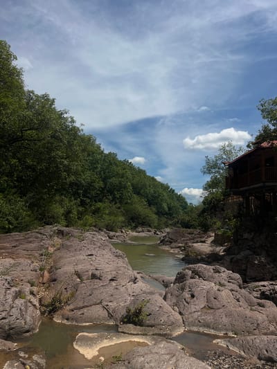 A serene landscape scene unfolds with a gently flowing river cutting through a rocky terrain. Lush green trees line the banks, creating a verdant backdrop. On the right side of the image, a rustic wooden structure, possibly a small house or cabin, is perched on stilts amidst the trees, its red-tiled roof adding a splash of color. The sky above is a dynamic canvas of blue with scattered white clouds. The scene appears to be located in Arambala, El Salvador, a region known for its natural beauty. The photograph captures a tranquil moment, inviting viewers to appreciate the harmonious blend of nature and rustic architecture.