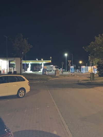 The image depicts a dimly lit outdoor scene at night, likely a rest stop or service area. A white station wagon is parked in the foreground, facing away from the camera. In the background, a gas station with illuminated pumps and a canopy is visible. Several large blue and white structures, possibly portable restrooms or waste disposal units, are also present in the midground. A portion of a truck is visible to the right of the gas station. The environment appears to be paved with concrete and asphalt, with some grassy areas and trees. The sky is dark and clear. The overall impression is that of a functional, utilitarian area, possibly along a highway or in a commercial zone, in Siemz-Niendorf, Germany.