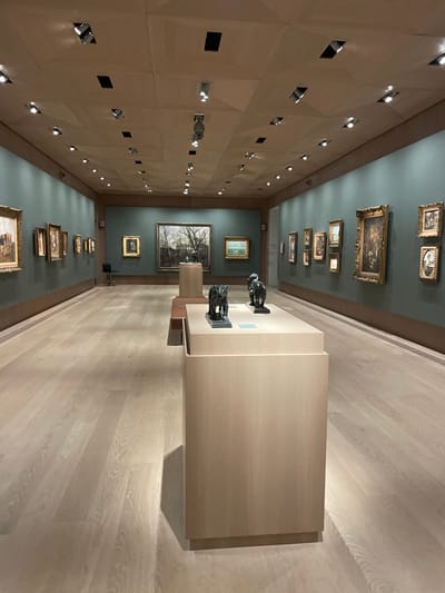 This image displays the interior of an art gallery, likely located within the Ordrupgaard Museum in Klampenborg, Denmark. The scene depicts a long, rectangular exhibition space with a light-colored wooden plank floor and muted green-gray walls. The ceiling is paneled with light-toned wooden geometric shapes, incorporating numerous recessed spotlights and dark rectangular apertures for lighting or ventilation.

Multiple framed paintings of various sizes, predominantly in ornate gold frames, are displayed on both side walls and the far end wall. The left wall exhibits a series of approximately nine paintings. The far wall features a large landscape painting depicting bare trees centered, flanked by smaller works; a smaller landscape painting is to the right of the central piece. The right wall showcases a collection of at least ten framed paintings, including portraits and genre scenes.

In the foreground and mid-ground, light-colored wooden pedestals are centrally positioned. A prominent, elongated display pedestal in the foreground supports two dark bronze sculptures, identifiable as large feline figures on dark bases. Behind this, a low wooden bench-like structure is visible. Further down the room, a smaller light-colored wooden pedestal holds a single dark bronze animal sculpture. A single black chair is situated against the far-left wall. No individuals are present within the gallery space. The lighting originates entirely from the ceiling fixtures, illuminating the artworks and the room uniformly.