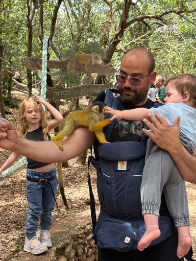 The image shows a man wearing glasses and a beard holding a toddler in a blue baby carrier. A yellow squirrel monkey is perched on the man's arm. A girl with blonde hair and blue jeans is standing below the monkey. The location appears to be an outdoor area with trees and rope. There is a sign with a monkey illustration and Hebrew writing, likely in Misgav Regional Council, Israel.