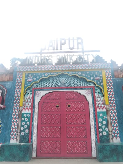 The image shows a colorful, ornate entrance to a building. A large, red door with a patterned surface and two gold knobs is centrally located. The door is flanked by vibrant murals depicting floral designs, peacocks, and a figure in traditional Indian attire. Above the door, a sign reads "INDIA'S PINK CITY" in large, white, three-dimensional letters. The overall style suggests a cultural attraction or themed establishment. The text "India's Pink City" likely refers to Jaipur, India, a city known for its distinctive pink architecture. However, the location provided is "Gudangkahuripan, Indonesia," which seems to be a mismatch based on the visual cues. The scene appears to be outdoors, with bright daylight casting a slight glare on the top of the image.