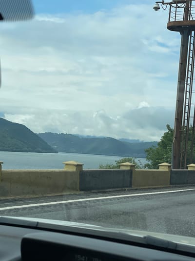 The image, oriented correctly, captures a daytime view from within a vehicle, showcasing a dramatic natural and infrastructural landscape.
In the upper part of the frame, a large metal gantry or bridge structure spans across, with a security camera or light fixture suspended from it. Below this structure, the sky is visible, featuring a mix of bright blue patches and soft, scattered white and grey clouds.
The central and lower portions of the image are dominated by a vast body of water, likely a river or a lake, stretching towards distant mountains. These mountains rise steeply from the water's edge, their dark green slopes receding into a hazy blue as they extend into the background. Trees and vegetation are sparsely visible on the near bank and more densely cover the mountains.
In the foreground, to the right, a section of a paved road with a distinct white line is present, bordered by a concrete barrier or railing that runs parallel to the water. The frame of the vehicle's interior, possibly a window pillar, partially occupies the far right side of the image. A partial digital display within the vehicle shows "1541 18°", likely indicating the time (3:41 PM) and an outdoor temperature (18 degrees Celsius).
The scene suggests a journey through a scenic area with significant geographical features and infrastructure, consistent with locations like the Iron Gates gorge near Drobeta-Turnu Severin, Romania. No specific human activity or people are depicted.