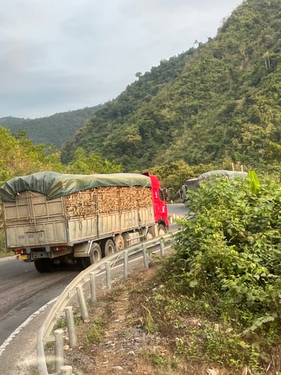 The image shows a truck on a winding road in Aê Thi, Vietnam. The truck is loaded with what appears to be a cargo of bamboo or similar material, partially covered by a tarp. The road is lined with a metal guardrail on one side. In the background, there is a steep, densely forested hillside. The weather seems overcast. Another truck is visible further up the road.