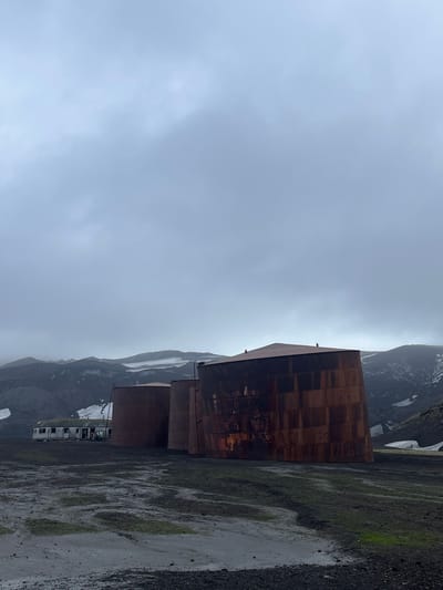 A cluster of weathered, rust-colored buildings stands against a stark, mountainous backdrop. The structures appear cuboid and cylindrical, displaying a patchwork of metal panels. Patches of snow cling to the rugged mountains. Below, the ground is wet, with puddles reflecting the overcast sky. The scene suggests a remote, industrial setting under gloomy weather conditions.