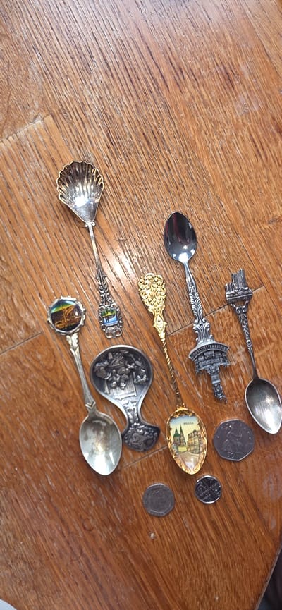 The image shows a collection of decorative souvenir spoons arranged on a wooden surface, likely a table. There are at least eight spoons visible, each with a unique design on the handle or the bowl. Some spoons depict landmarks or scenes, such as a seashell, a cityscape with the word "PRAGUE" visible, and a building resembling a castle or tower. There are also two coins visible on the wooden surface, one with the numeral "50" and another with a cross pattern. The wooden surface has a prominent grain and appears to be an indoor setting. There are no people or animals present in the image. The city of Marum, Netherlands, is not depicted or implied in this image.