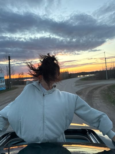 This image, captured in Dubnik, Russia, portrays a person leaning out of a car window, their back mostly to the camera. The individual has dark, curly hair styled in a messy bun and is wearing a light-colored, possibly grey or light blue, zippered hooded jacket. Their gaze appears directed towards the distant horizon, where the sun is setting or rising, casting warm orange and pink hues across the sky. Above this colorful band, the sky transitions to cooler tones of grey and blue, filled with scattered clouds.
The setting appears rural or semi-rural, featuring a dirt or unpaved road winding into the distance, flanked by bare trees and utility poles. Part of a sign with partial Cyrillic text is visible in the lower left foreground. The scene conveys a sense of freedom or quiet contemplation, set against a beautiful twilight landscape. The overall composition is dynamic, with the horizon appearing tilted.