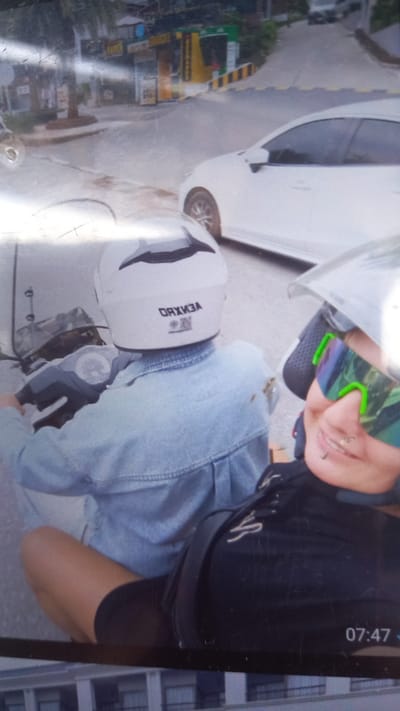 A low-angle, slightly blurred photograph shows two individuals on a motorcycle. The time 07:47 is visible in the upper right corner. A white vehicle is parked to the left. The person in front wears reflective sunglasses and a helmet. The person behind wears a white helmet and a denim jacket. The letters "VENXO" are visible on the back of the helmet. The location appears to be an urban street in Ban Tak Daet, Thailand. Buildings and signs are visible in the background, along with street markings.