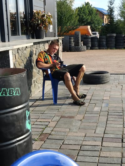 The image depicts a man sitting on a blue plastic chair outdoors, with a cigarette in his mouth, looking at his phone. He is wearing a colorful, patterned short-sleeved shirt and black shorts. His legs are bare and he is wearing dark sandals. In the foreground to the left, a large black barrel with green text and a green graphic of an animal is visible. Behind the man, a large black tire is lying on the ground. In the background, stacks of tires are arranged, a dump truck, and a building with a blue roof can be seen. The ground is paved with rectangular bricks. The scene appears to be in an outdoor industrial or storage area, possibly a tire shop or a junkyard. The presence of the building with a blue roof and the general landscape suggests a location in a city or town in Ukraine, potentially in the city of Novoselytsia.
