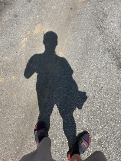 The image shows a shadow of a person standing on asphalt. The shadow is cast by the sun, indicating it is daytime. The person is wearing sandals. A thin, light blue object is visible on the asphalt to the right of the shadow. The ground appears to be a road or parking lot, with faint yellow lines visible. The location is the city of Lebanon, United States of America.