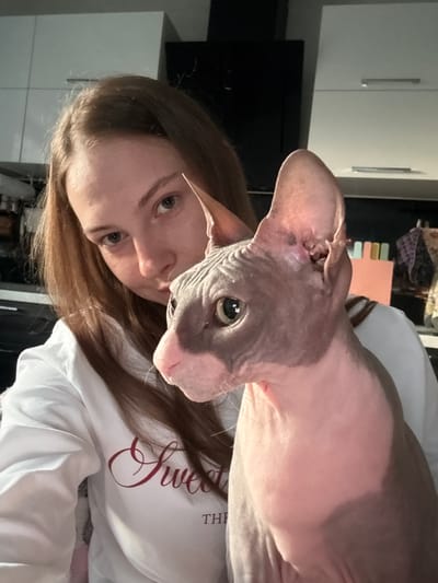A selfie is taken by a young woman with long brown hair, wearing a white sweatshirt with red lettering. She is holding a hairless cat, a Sphynx breed, which has pink and gray coloring and large ears. The cat is looking to its right, while the woman looks at the camera. They are indoors, with kitchen cabinets visible in the background. The location is identified as Novaya Ivanovka, Russia.