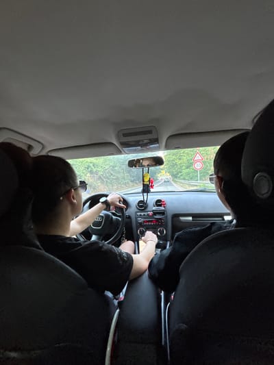 The image is taken from the back seat of a car driving on a road. Two people are visible from behind. The person in the driver's seat is wearing sunglasses and driving the car. The person in the passenger seat is partially visible. A speed limit sign of 30 km/h is visible outside the car on the right side of the road. The road appears to be in a rural or forested area. The scene is consistent with being on the road leading to or from Kresna, Bulgaria, known for its scenic mountain roads.