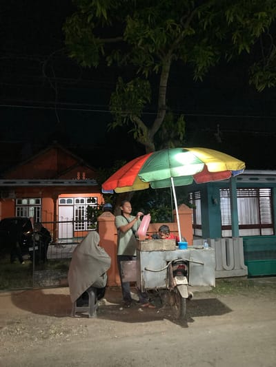 An overhead shot captures a nighttime scene on a street in Proyonangg, Indonesia. The foreground is dominated by a dirt and gravel road. To the left, a dark-colored car is partially visible. In the center, a person wearing a grey headscarf sits on a dark grey stool, facing away from the camera. Beside them, a food cart, attached to a motorcycle, is illuminated by the colorful canopy of a multi-colored umbrella above it. A man stands behind the cart, holding a pink plastic bag, and a young child in a red shirt is seated on the cart. The background features two houses: one on the left is orange with white window frames, and the one on the right is teal with white-framed windows and visible curtains. The sky is dark, with a large tree casting shadows across the scene. The license plate of the motorcycle is visible and reads "S702 09".