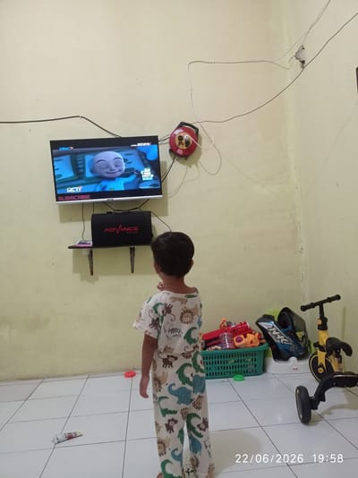 A young child stands in a room with light yellow walls, facing a television that displays a cartoon character. The child is wearing pajamas with an animal print. A black speaker with "ADVANCE DIGITALS" branding is mounted on a shelf below the television. To the right of the television, a red extension cord reel hangs on the wall. On the floor, a green plastic basket is filled with toys, including a toy gun and a helmet. A yellow and black tricycle is also visible. The floor is tiled. A timestamp in the lower right corner reads "22/06/2026 19:58". This scene is consistent with an indoor residential setting in Sukaraja, Indonesia.