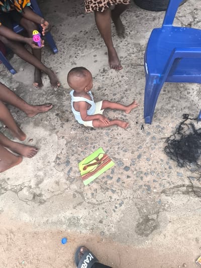 The scene is outdoors, likely a residential area in Abraka, Nigeria, as indicated by the presence of bare feet and the informal arrangement of people and objects. A baby is sitting on a concrete surface, wearing a light-colored undershirt and shorts. Several adults and children are present around the baby, evidenced by the presence of multiple bare feet. A blue plastic chair is positioned nearby, along with a piece of synthetic hair. A colorful mat is located near the baby. A blue plastic bottle cap and a single sandal are at the bottom of the frame. A child is holding a pink and purple toy.
