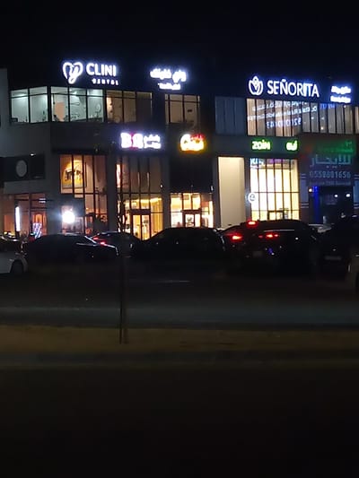 The image captures a vibrant commercial street scene at night in Al `Aziziyah, Saudi Arabia. Multiple businesses are housed in modern buildings with large glass windows, all brightly illuminated against the dark sky.

In the upper left, a prominent neon sign displays "CLINI DENTAL" with a heart-shaped logo, indicating a dental clinic. To its right, another sign shows "SENORITA" with a floral or leaf motif. Below "CLINI DENTAL," a "BR" sign, possibly for Baskin Robbins, is visible. Underneath "SENORITA," a green neon sign reads "ZOIN." Further to the right, a sign features Arabic text that translates to "للايجار" (For Rent) along with a phone number, "0558801656." Other indistinguishable Arabic script also appears on several signs.

The street level is bustling with several cars, mostly dark-colored sedans and SUVs, either parked or moving slowly. Their brake lights and reflections add to the dynamic nighttime glow. Interior lights of the businesses are clearly visible through their windows, suggesting they are open and active. A dark street pole stands in the foreground, partially obscuring the view of the vehicles. The overall atmosphere is one of active commerce and urban life after dark, typical of a shopping or business district.
