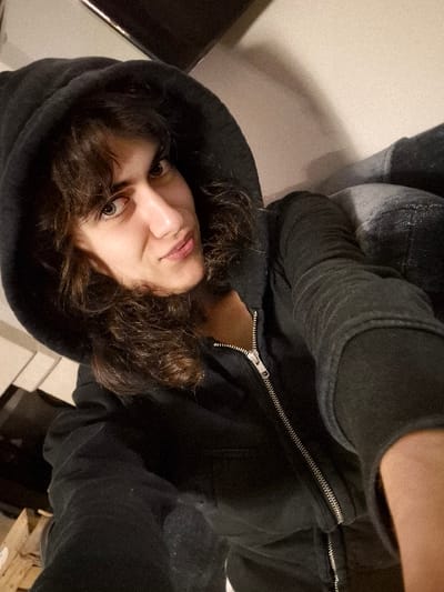 An indoor selfie captures a single individual in Châtelaillon-Plage, France. The subject is a person with medium-length, wavy dark brown hair, wearing a dark (likely black) hooded sweatshirt or jacket with the hood drawn over their head. A silver-colored zipper runs down the front of the garment and is partially unzipped. The individual has light-colored eyes, prominent eyebrows, and is looking directly into the camera with a subtle, closed-lip smile. The lighting appears to be artificial and warm. In the background, a light-colored wall is visible. To the upper left, a dark, rectangular, reflective object, possibly a television screen or monitor, is partially visible. In the lower left corner, stacked wooden objects, resembling pallets or lumber, are partially visible. To the right of the subject, a dark, textured object, potentially a piece of furniture, is partially visible. The subject's right arm (viewer's left) is extended, holding the camera, indicating a self-portrait.