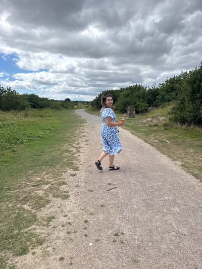 A young woman with dark hair tied back walks on a gravel path in a park. She wears a blue and white floral dress and black sandals. She holds an orange can in her right hand and looks back over her left shoulder, smiling. The path is lined with green grass and bushes. In the background, there are trees and a large rock formation. The sky is partly cloudy with scattered white and gray clouds. The location is the Blean Woods National Nature Reserve in the Borough of Swale, United Kingdom.