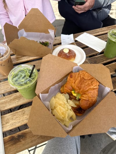An elevated view captures an outdoor dining scene on a slatted wooden table, likely in Chula Vista, United States. The foreground features an open kraft paper takeout box containing a large croissant sandwich filled with what appears to be egg and greens, alongside a portion of plain potato chips. To the left, a clear plastic cup with a black straw holds an iced matcha latte, displaying ice cubes and a foamy green beverage. Next to it, partially visible, is another clear plastic cup containing iced coffee.
Further back on the table, another open kraft paper box contains a smaller portion of mixed greens or microgreens. A white paper plate holds a sugar-dusted donut topped with a small dollop of white cream or butter. A crumpled white receipt and a folded white napkin are also present on the table.
Two individuals are partially visible: a person in the upper left wears a light pink collared shirt, and another person in the upper right wears a dark plaid shirt over dark trousers, holding a dark-colored smartphone with a green case in their right hand. A second iced matcha latte in a clear plastic cup is visible in the upper right corner. The lighting suggests an outdoor setting under natural light, and the chairs beneath the table are dark-colored.
