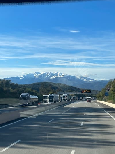 The image presents a daytime view from inside a vehicle on a multi-lane highway, likely the A9 autoroute near Le Perthus, France, facing generally south towards the Pyrenees mountains.
The sky is predominantly clear blue with scattered wispy white clouds and a distinct, elongated lenticular cloud centered in the upper frame. In the background, a majestic range of snow-capped mountains, consistent with the Pyrenees, stretches across the horizon, flanked by forested hillsides.
The highway features at least three lanes in the direction of travel, separated by a concrete median barrier. The leftmost lane appears relatively clear. The middle and right lanes exhibit significant traffic congestion, primarily composed of commercial semi-trucks. Visible trucks include a silver tanker truck in the left-center of the foreground and numerous white tractor-trailer units extending into the distance. A red passenger vehicle is discernible amidst the truck traffic further ahead in the middle lane.
Overhead, a gantry sign spans the highway, displaying amber text and an orange warning icon, likely indicating traffic congestion or advisories. On the right shoulder, a blue directional sign shows "10 km" and an arrow pointing right, with smaller text below, possibly indicating a route or destination. A triangular warning sign is positioned beneath it. A small red sign is visible on the median barrier in the foreground, possibly indicating a temporary stop or warning. Bright sunlight casts long shadows of vehicles and roadside structures across the asphalt pavement. No individuals are clearly visible.