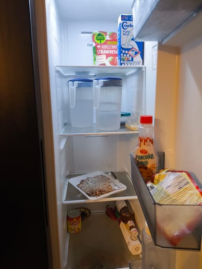 The image shows an open refrigerator, likely in a home kitchen in Imus, Philippines. The refrigerator is well-lit, suggesting it is daytime or the interior light is on. Various food and drink items are visible on the shelves.

On the top shelf, there are two boxes of Happy Village Organic Gently Dried Strawberries and a carton of Cow & Gate Pure Milk. Next to the milk is a white box with text that appears to be related to appliance information.

The middle shelf holds two large, clear pitchers filled with water and blue lids. To the right of the pitchers, there is a bottle of Clara Ole Pancake Syrup with a red cap, and a small plastic bag containing some food items.

On the bottom shelf, a white plastic tray holds a portion of ground meat or shredded meat. To the left of the tray is a can of Alaska Evaporada. In the door compartment, a dark bottle with a brown cap and a white box with yellow accents are visible, along with some red chili peppers. A clear plastic drawer at the bottom contains packaged food items, including one with visible "Nutrition Facts" and "Ingredients" text, mentioning "Mama Sita's Oyster Sauce."

The scene is one of a stocked refrigerator, with a variety of common household food items. There is no indication of people or specific events, but the presence of staple goods suggests it's a typical day in a residential kitchen.