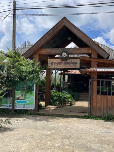 A wooden building with a pitched roof, decorated with a Christmas wreath, is pictured in Thôn 4, Vietnam. The sign above the entrance reads "B'nom Kitchen". A wooden walkway leads into the building, which appears to be a restaurant or cafe. Lush greenery surrounds the structure, with trees and plants visible in the foreground and background. A cloudy sky with patches of blue can be seen above. To the left of the building, a poster displays an image of a house by a pond. Electrical wires and a utility pole are also present in the scene. The ground in front of the building is a mixture of dirt and gravel.