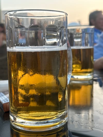 In Plataniás, Greece, two glasses of beer are on a table, with the foreground glass being the most prominent. This glass is filled with golden beer and topped with a foamy white head, condensation visible on the outside. Behind it, another glass of beer is partially visible. To the left of the foreground glass, a pack of cigarettes is on the table. The background is out of focus, showing a blurred figure of a person wearing a blue shirt. The lighting suggests it is late afternoon or early evening, with a warm, soft glow in the sky. The overall scene depicts a relaxed, outdoor setting, possibly a taverna or bar near the coast, typical of the Plataniás region.