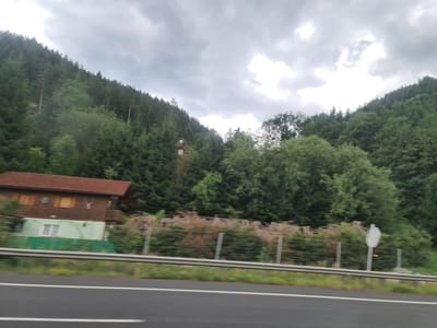 The image captures a roadside view in Leoben, Austria, on a cloudy day. In the foreground, a dark asphalt road with a white line runs horizontally across the frame. A metal guardrail separates the road from a grassy embankment.
Behind the embankment, a low green structure with a white-framed window is visible, attached to a larger wooden building with a reddish-brown roof. This wooden building has multiple windows with what appear to be wooden shutters. A decorative element, possibly a set of antlers, is mounted on the facade of the wooden building. A fence made of metal posts and wire mesh runs along the property line, bordering some dry, brush-like vegetation and more green trees.
The background is dominated by a dense, dark green forest that climbs a steep hillside. The trees are varied in height and type, creating a thick canopy. The sky is overcast with heavy, grey clouds, suggesting a high probability of rain. No people or vehicles are clearly discernible in the image.