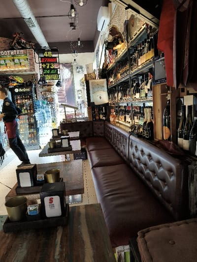 This image depicts the interior of a commercial establishment, likely a cafe, bar, or bottle shop, located in Riccione, Italy. The space features a long, narrow layout with a functional, rustic-industrial aesthetic.
On the left side, a staff member is visible from the mid-torso upwards, wearing a dark long-sleeved shirt and a red apron, standing adjacent to a glass-front beverage refrigerator stocked with various bottled and canned drinks, including "Warsteiner" beer. Above this area, a "SMOOTHIES" sign, a yellow helmet labeled "GAME", and a red model motorcycle are mounted on a faux brick wall. A prominent black sign displays "46", "27 BIAGIO", "+73", "24 ALE", and "IN FUEL". The floor is composed of light-colored tiles.
The central area is furnished with several rectangular tables featuring distressed wooden tops. Each table is set with a black tray containing a black napkin dispenser, some bearing a "USSE CAFE" logo, alongside one or two golden-colored metal cups and sugar packets. Parallel to these tables, along the right wall, runs an extensive L-shaped continuous bench upholstered in brown, button-tufted leather or faux leather.
The right wall is dominated by multiple tiers of metal shelving densely filled with a wide assortment of wine and spirit bottles. Interspersed among the merchandise are decorative elements including an American flag design, framed pictures, and a "WELCOME" sign. Overhead, multiple industrial-style pendant lights with exposed bulbs hang from a dark ceiling, which also features an exposed metal air duct and a white wall-mounted air conditioning unit. Additional bottles are displayed on a low counter surface in front of the bench. A partial view of another brown upholstered seating unit is visible in the extreme right foreground.
One individual, identified as a staff member, is present and standing. No direct interactions or specific actions beyond standing are discernible. The environment is well-lit and appears organized.