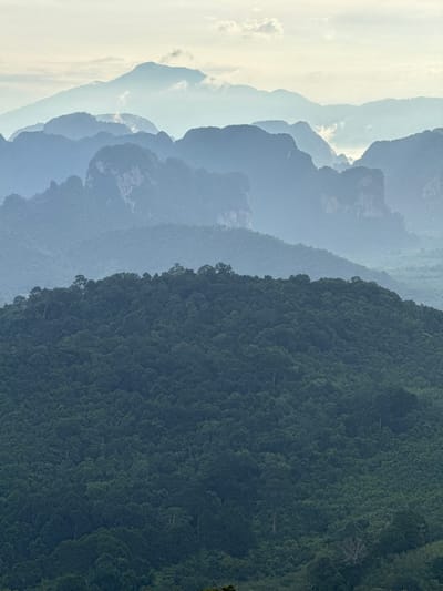 The image depicts a layered mountain landscape, likely from a high vantage point. In the foreground, a dense, dark green forest covers a rolling hill. Beyond this, successive layers of mountains recede into the distance, becoming progressively lighter and more obscured by atmospheric haze. The farthest mountains are mere silhouettes against a pale sky with wispy clouds. The overall impression is one of natural grandeur and tranquility. The location is identified as Ban Din Daeng Nui, Thailand.
