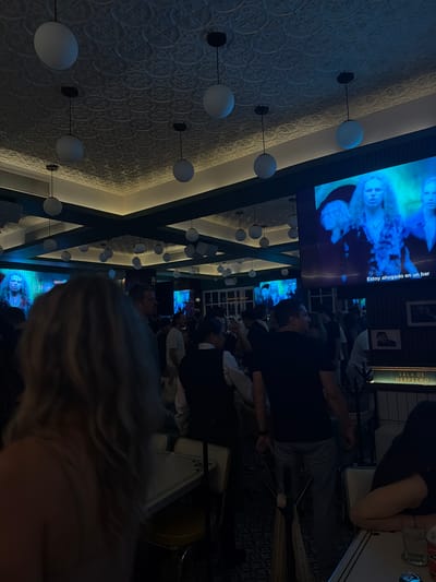 The photograph captures the interior of a bar, likely located in Puerto Vallarta, Mexico, as suggested by the Spanish phrase "Estoy ahogado en un bar" visible on one of the television screens. The bar's decor includes textured walls, spherical lighting fixtures, and dark-painted structural elements forming an intersecting pattern across the ceiling. Several televisions are mounted along the walls, displaying digital images.
Patrons are present in the scene, engaged in casual social interactions, such as watching television and talking. The foreground is somewhat obscured by a blurry figure with blonde hair. A table with glasses and other objects can be seen. The overall atmosphere is dimly lit.