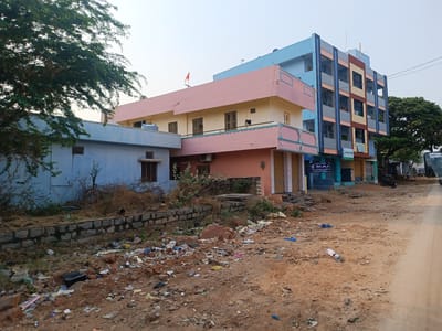 The scene is a street in Kothacheruvu, India. There are several buildings along the street, painted in various colors. One building is a light blue, while another is a combination of pink and yellow. A third building is a taller, multi-story structure with blue, gray, and brown sections. The street and adjacent areas are littered with trash and debris. Vegetation is visible near the buildings and along the street. The sky is overcast. A tree is on the left edge of the photo, partially obscuring the blue building. A street is visible on the right side of the image.