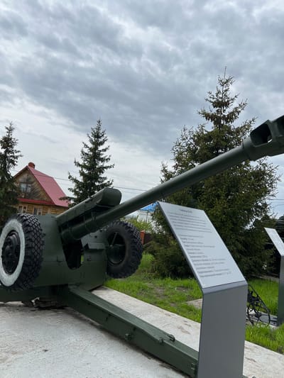 The image shows a Soviet-era 122mm D-30 howitzer on display outdoors. The howitzer is painted green and has two wheels with white rims. A sign next to the howitzer provides information about its specifications, including its caliber, type, crew size, weight, range, rate of fire, and vertical aiming angles. The howitzer is situated on a concrete slab. In the background, there are evergreen trees, a cloudy sky, and a wooden building with a red roof. The sign indicates that the howitzer is located in Введенская Слобода, Russia.