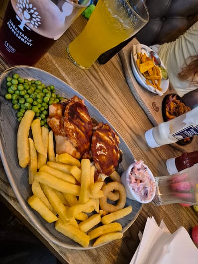 The image depicts a table at a restaurant in the Breckland District, United Kingdom. A grey, patterned plate is centrally positioned, containing two glazed pies, a portion of garden peas, a serving of French fries, onion rings, and a small bowl of coleslaw. Adjacent to the plate are two glasses; one filled with an orange-colored beverage and the other with a dark liquid, the latter displaying the text "UNBEATABLE REFRESHMENT." The table also holds a wooden platter with a small dish of tortilla chips and accompanying dips, a small black frying pan with sauteed vegetables, a bottle of mayonnaise, a bottle of ketchup, a glass of water with fruit slices, and a stack of white napkins. Part of a person is visible in the upper left corner. The table's surface is made of light-colored wood.