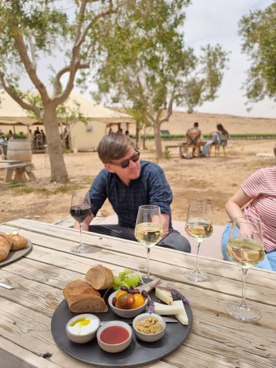 **Scene:** An outdoor wine tasting at a vineyard.

**Location:** Mizpe Ramon, Israel.

**People:** A man in a blue plaid shirt and sunglasses is seated at a wooden table. Another person wearing a red and white striped shirt and blue pants is at the same table. A group of people in the background are sitting in chairs, possibly also partaking in the wine tasting.

**Objects:** There are several wine glasses on the table, some containing white wine and one containing red wine. A black plate holds a cheese board arrangement, consisting of cheese slices, fruits (grapes, tomatoes), bread slices, and various dips in small bowls. There is an additional plate of bread, partly visible, at the edge of the table. A tree is to the left of the man, and tents and wine barrels are visible in the background.

**Actions:** The man is seated at the table, apparently involved in the wine tasting. The people in the background are seated and possibly engaging in conversation or other activities related to the vineyard experience.

**Interactions:** The people at the table are seemingly interacting, enjoying the wine tasting and the accompanying food.

**Environment:** The location appears to be a vineyard with outdoor seating under trees, creating a relaxed and casual atmosphere. The background shows desert terrain, characteristic of the Mizpe Ramon region.