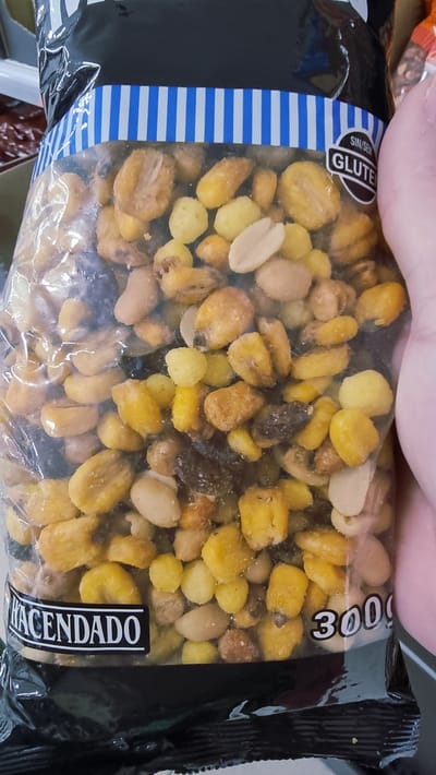 The image shows a close-up of a bag of mixed nuts and corn snacks. The bag is mostly black with a blue and white striped band across the top. A "SIN/SEM GLUTEN" (without gluten) label is visible on the right side of the bag. The brand name "HACENDADO" is printed in white on a black label at the bottom left of the bag. The weight "300g" is visible in white text on the right side of the bag. The snacks appear to be roasted corn kernels, peanuts, and possibly other small fried or roasted items. A person's hand is visible on the right side of the image, holding the bag. The background is blurred and does not provide any identifiable environmental context related to the city of Olot, Spain.