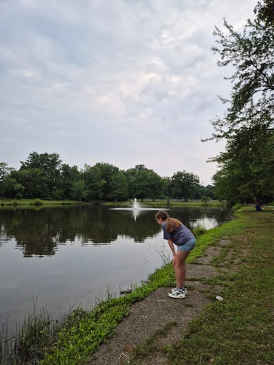 A young girl with long brown hair is bending over near the edge of a pond, looking into the water. She is wearing a blue t-shirt and denim shorts. The pond is surrounded by lush green trees and grass. In the background, a fountain sprays water into the air. The sky is overcast with gray clouds. The scene appears to be a park in South River, New Jersey, United States of America.