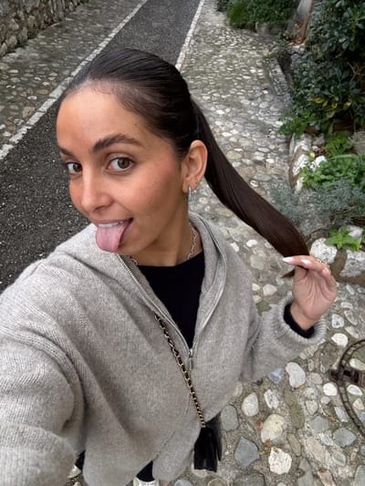 A close-up, high-angle selfie captures a female individual standing on a cobblestone pathway within the city of Saint-Paul-de-Vence, France. The subject, with medium skin tone and dark brown hair pulled into a high ponytail, looks directly at the camera with a playful expression, her tongue extended. She is wearing a light grey knitted zip-up hooded cardigan over a black top, accessorized with a silver-colored chain necklace and a black small shoulder bag featuring a gold chain strap. Her left ear displays a small hoop earring with clear stones on the lobe and a small stud on the upper cartilage. Her right hand, with light-colored manicured fingernails, is visible holding a section of her ponytail. The background features an irregular cobblestone path with varying shades of grey and beige, containing a darker, smoother central strip. Green foliage and a partial stone wall border the path on the right side. The scene is illuminated by diffuse natural light.