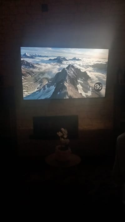 The image shows a television screen displaying a stunning landscape of snow-capped mountains partially obscured by clouds. A circular logo, featuring stylized letters that appear to be "BG" or a similar monogram, is visible in the upper center of the screen. To the right of the television, a dark surface, possibly a table, holds a decorative item that resembles a small potted plant with white flowers. The scene is dimly lit, suggesting it is either nighttime or the room has its lights off, with the television being the primary light source. The overall atmosphere is calm and serene, with the focus on the majestic mountain vista. The television is positioned against a textured wall, possibly brick or stone, which is also dimly illuminated. The location context provided is Oliveira do Bairro, Portugal.