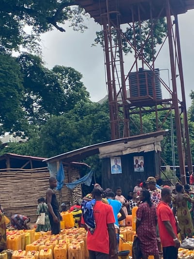 The image depicts a scene in Nguzu Edda, Nigeria, showing a group of people lined up near a water tower or elevated tank structure. The crowd appears to be waiting, possibly to collect water. Many yellow jerrycans are stacked nearby, likely for transporting water. The people are wearing various clothing styles, and some are carrying items. There are political posters on a structure adjacent to the waiting line. The surroundings consist of trees and a building of local architecture, suggesting a rural or semi-urban environment.