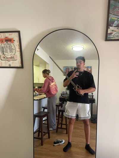 An indoor residential scene in Arlington, United States, is depicted through a large, arched, black-framed mirror. In the foreground reflection, a male individual, appearing to be in his twenties with dark, curly hair and facial hair, is visible from the upper thigh upwards. He wears a black short-sleeved t-shirt, white athletic shorts with black side stripes, and black socks with white Nike swooshes. He holds a black smartphone in his left hand, obscuring part of his face, and an ornate, antique-style sword with a brass hilt in his right hand, with the blade angled upwards and left. On the light brown wood-look floor, a black Converse-style sneaker with white laces is present near his feet.
In the background reflection, a female individual, also appearing to be in her twenties, with light brown hair secured by a claw clip, stands at a dark kitchen countertop. She wears a pink hooded sweatshirt with white lettering on the back and grey wide-leg pants, appearing to be engaged in an activity involving items on the counter. The kitchen area features light-colored cabinets and two dark wooden bar stools. A circular ceiling light fixture is reflected above. Behind the male's reflection, an abstract, colorful artwork hangs above a dark console table with various objects.
Outside the mirror's reflection, on a plain, light-colored wall, two framed items are visible: To the left, a "1979 ALBUQUERQUE LASERS Home Matches WORLD CLASS VOLLEYBALL" poster. To the right, a partially visible framed picture, possibly of baseball cards.